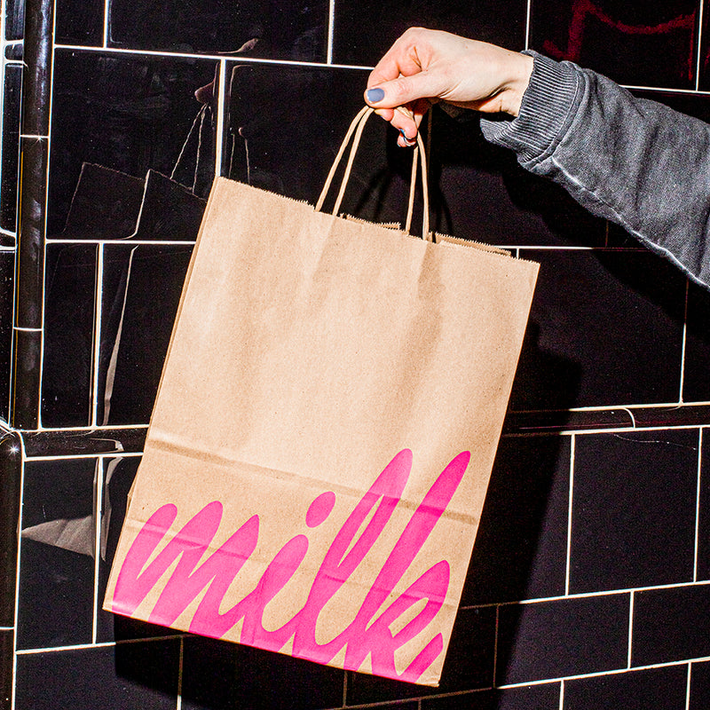A front view of a hand holding a branded Milk Bar kraft bag against a tiled wall.
