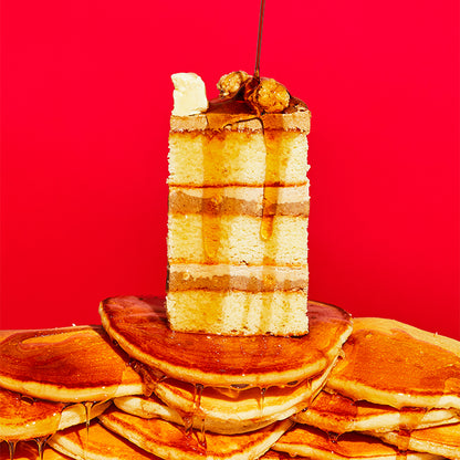 A slice of pancake cake sitting on a wide stack of pancakes with maple syrup drizzled on top 