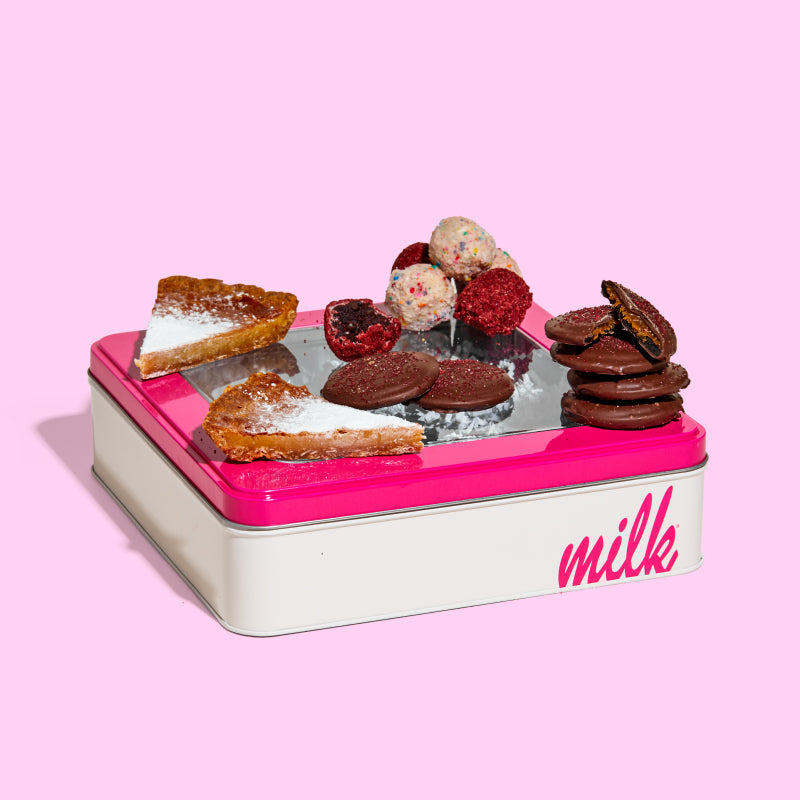 Side view of a milk bar branded tin with 2 milk bar pie slices, 6 mini chocolate-covered strawberry snaps, and assorted b'day and red velvet cheesecake cake truffles sitting on top.