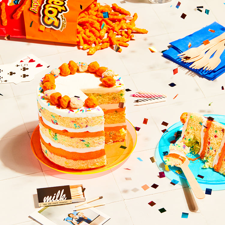 A decorative table spread containing an opened bag of Crunchy Cheetos spilling out of the bag beside a whole Cheetos x Milk Bar Birthday Cake with a slide removed. 