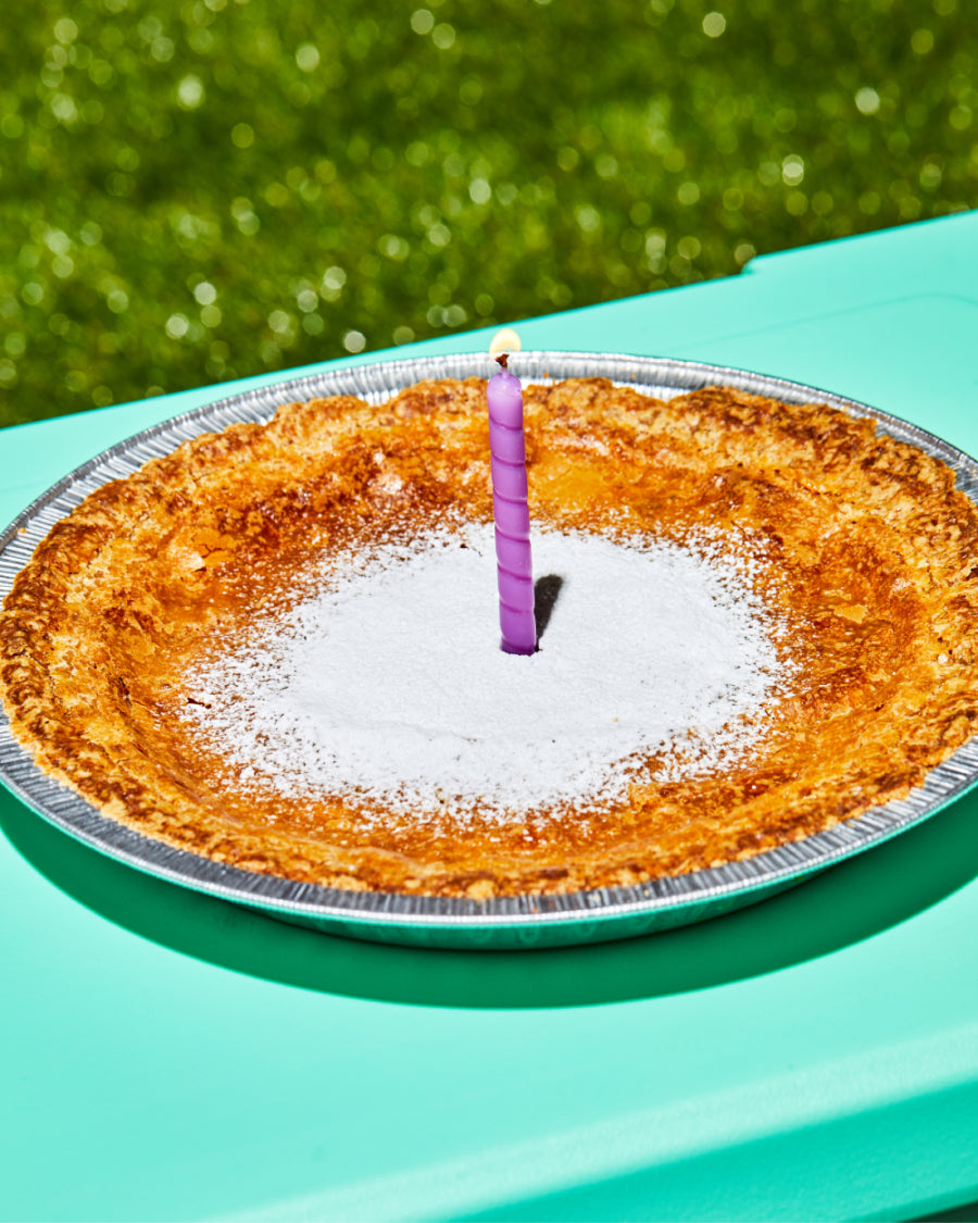 A whole milk bar pie with a single candle in the middle, sitting on a table outside on a beautiful sunny day
