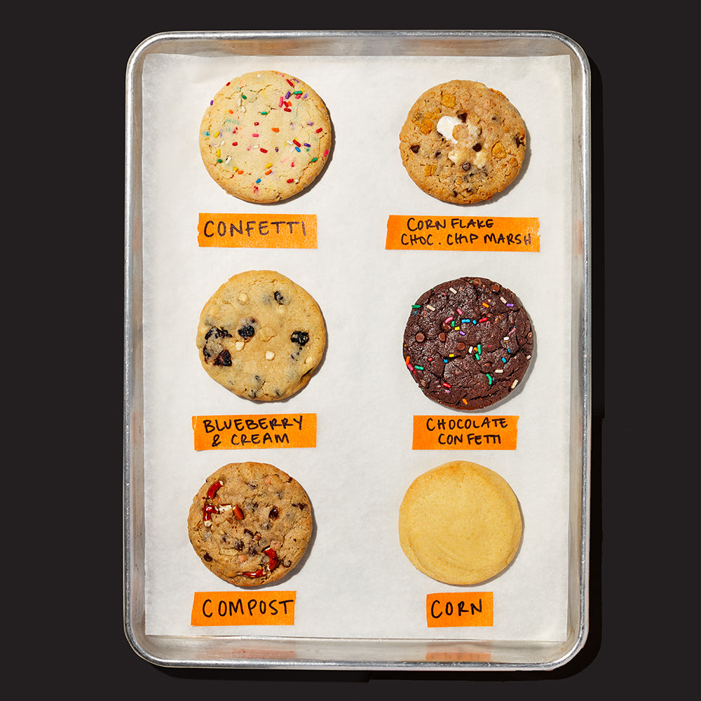 overhead of a baking tray sheet with 6 assorted cookies, each with a name label below in orange tape