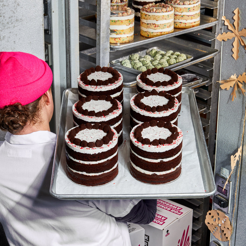 A baker carrying a tray of freshly baked Peppermint Bark Cakes into a walk-in fridge.