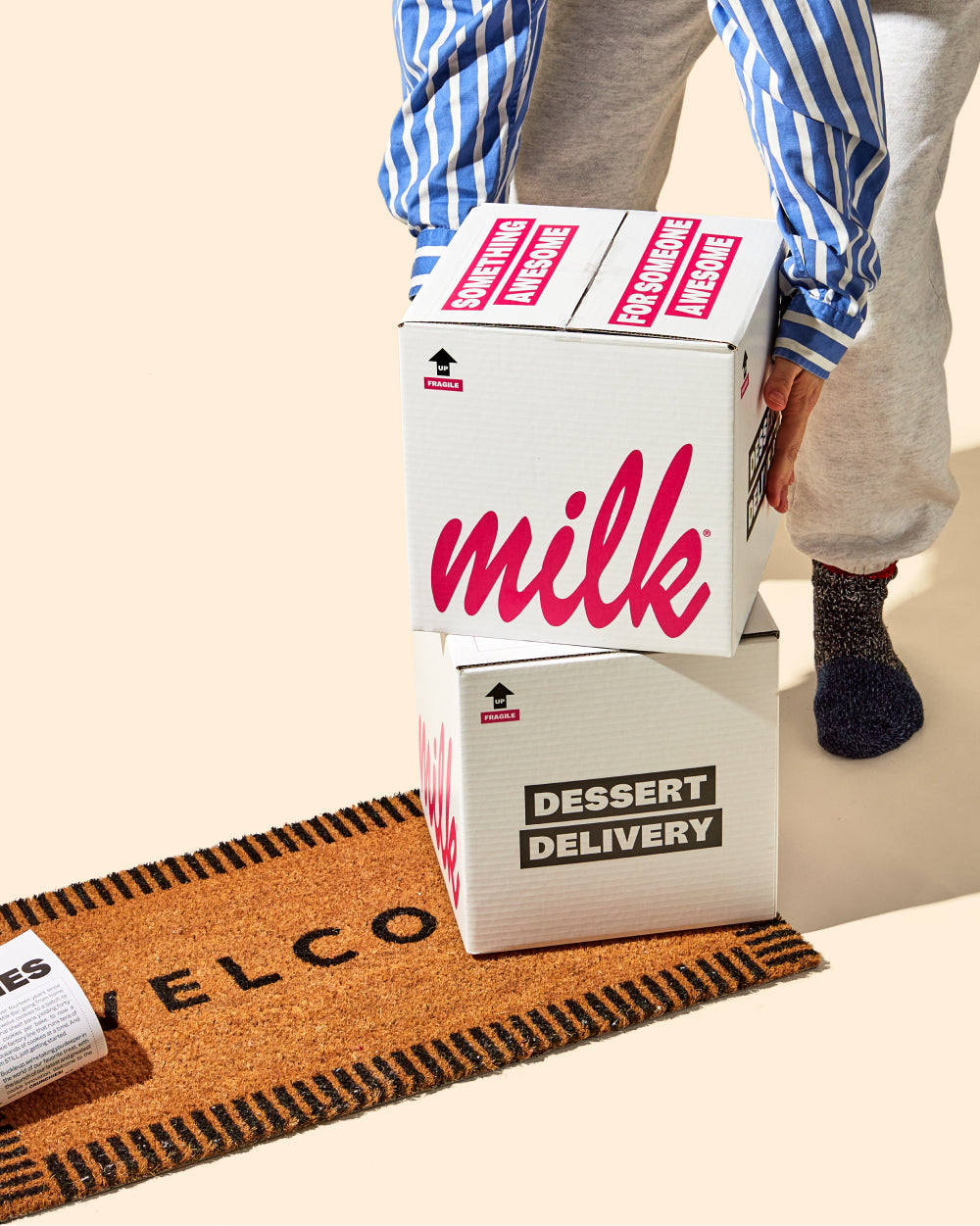 A person grabbing 2 stacked branded Milk Bar delivery boxes from their welcome doormat.