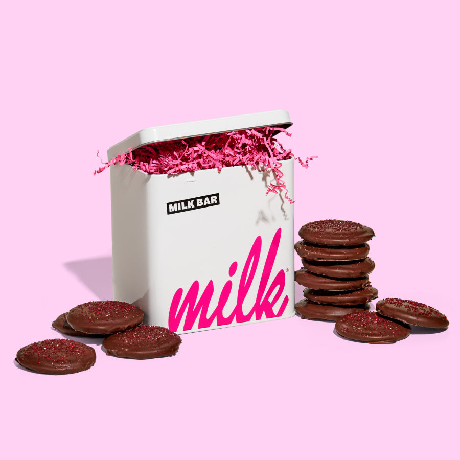 A milk bar branded tin beside two stacks of mini chocolate covered strawberry snap cookies