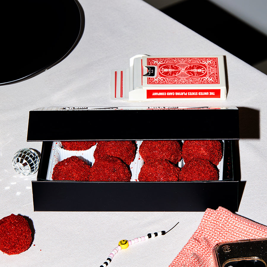 Side view of a red velvet cheesecake truffle dozen box with the lid ajar on a white tablecloth surrounded by decorations and a deck of playing cards.