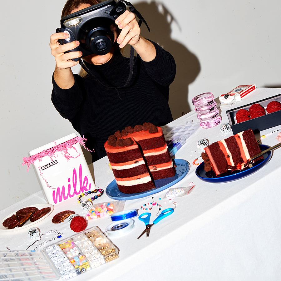 A photographer taking a picture of a dessert table containing a whole red velvet cheesecake, mini chocolate covered strawberry snap cookie, and red velvet cheesecake truffles