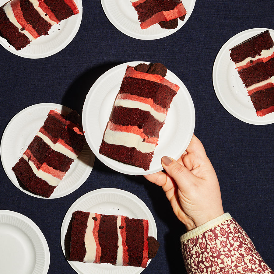 overhead of a dessert table containing several plates of Red Velvet Cake Slices, with a human hand holding up one plate 