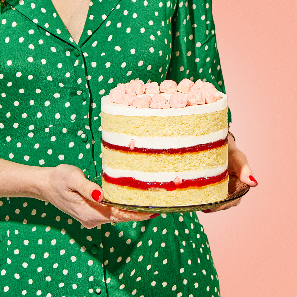 A close up view of a person in a polka dot dress carrying a 6 inch strawberry shortcake cake on a glass plate