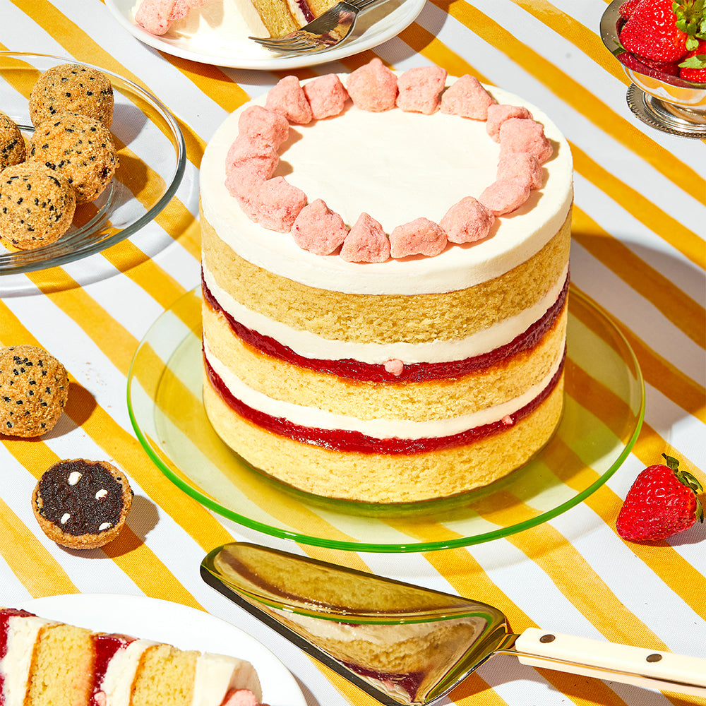 A 6 inch strawberry shortcake cake on a festive outdoor table, surrounded by s'mores cake truffles, fresh strawberries, and a cake slice serving utensil. 