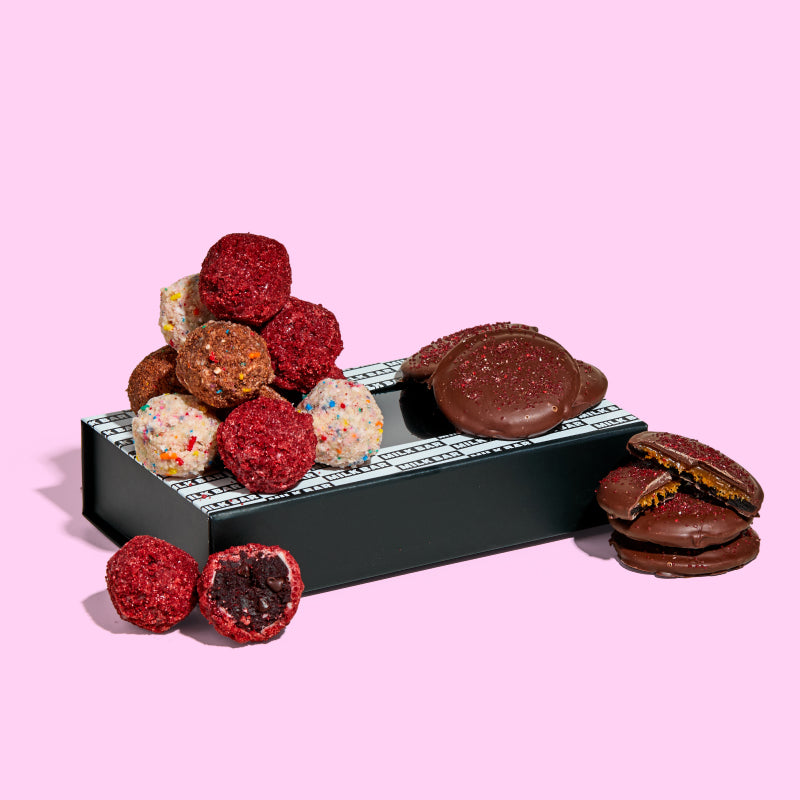 A pile of assorted cake truffles and a stack of limited-edition chocolate covered strawberry snap cookies sitting on a giftable branded box.