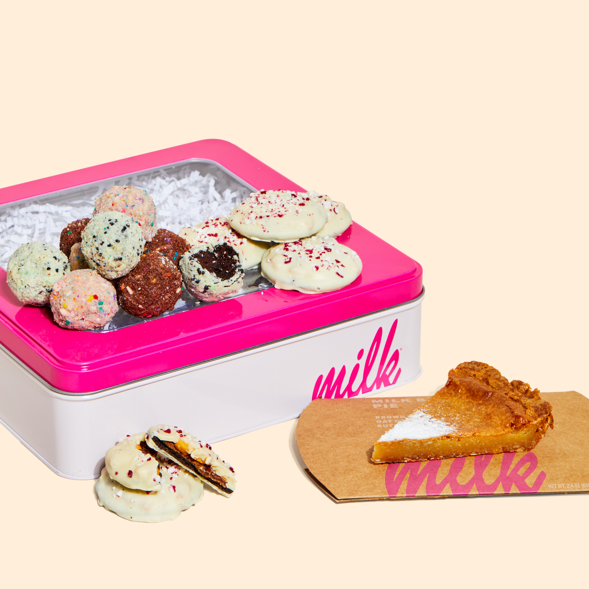 A pile of assorted truffles and mini peppermint bark snap cookies sitting on top of a milk bar branded tin, with a slice of Milk Bar pie on the side.